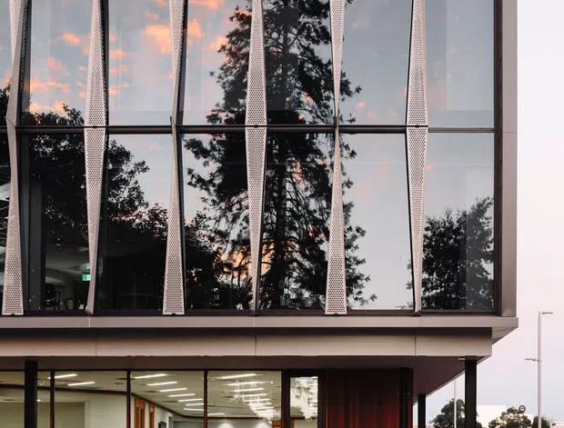 Exterior view of a contemporary multi-storey commercial building with a highly glazed upper facade, dark framing, and repeated perforated vertical screens. The ground floor contains a brightly lit office or commercial interior visible through glazing, with warm timber cladding at the entrance. The building fronts a marked asphalt parking area and appears in an urban setting at dusk, with trees and reflected sunset light in the glazing.