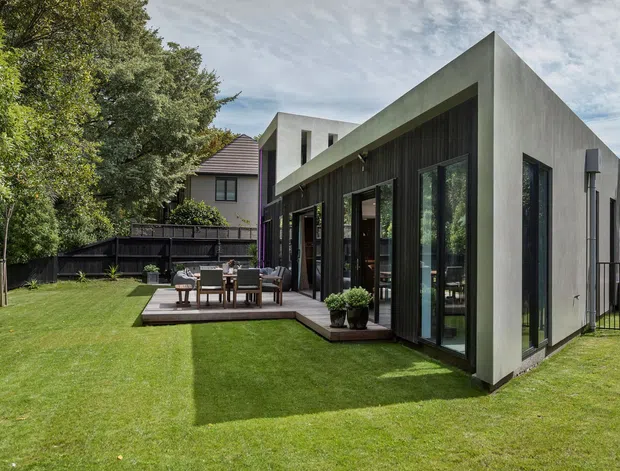 Contemporary single-storey extension with a sharply angled roofline, smooth grey rendered walls, dark vertical timber cladding and extensive black-framed glazing. The building opens onto a timber deck with an outdoor dining setting, surrounded by a landscaped lawn, mature trees and fencing.