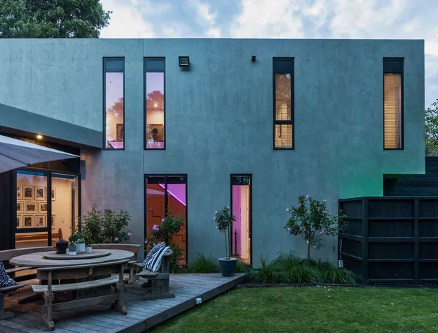 Contemporary two-storey residence with a simple flat-roofed rendered facade, narrow vertical windows, and dark-framed glazing. The home opens onto a timber deck and landscaped lawn, with an outdoor dining area beneath a large umbrella. Warm interior lighting and colourful reflections are visible through the windows and glazed doors, while a dark screened structure extends along the right side.