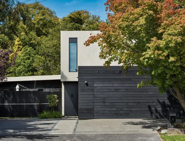 Street-facing exterior of a contemporary multi-level residence, featuring a flat-roofed white rendered upper volume, a dark horizontal timber-clad garage and entry frontage, narrow vertical glazing, and a concrete driveway. Mature trees and landscaped planting frame the facade.