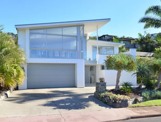 Bright exterior view of a contemporary multi-level coastal-style home with white horizontal cladding, expansive upper-level glazing, a flat projecting roof, integrated double garage, balcony, and landscaped front garden with palms and stone-edged planting beds.