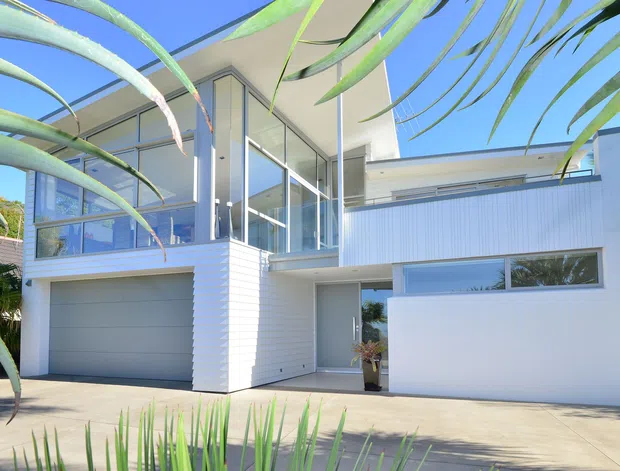 Bright exterior view of a contemporary multi-level residence with crisp white cladding, expansive glazed corner windows, a projecting upper volume, and an integrated double garage. Tropical foliage frames the foreground, while a broad concrete driveway leads to the entry and garage.