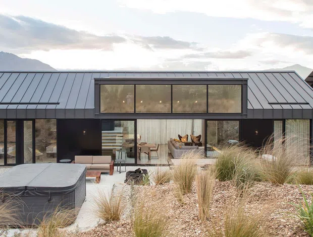 A contemporary single-storey residence with a dark metal-clad facade and low-pitched roof, viewed from a landscaped outdoor area. Large glazed openings reveal an open-plan living and dining space, while a raised central clerestory-like window band brings light into the interior. The foreground features naturalistic grasses, gravel landscaping, and a dark built-in spa or water feature. Mountainous terrain is visible in the background.