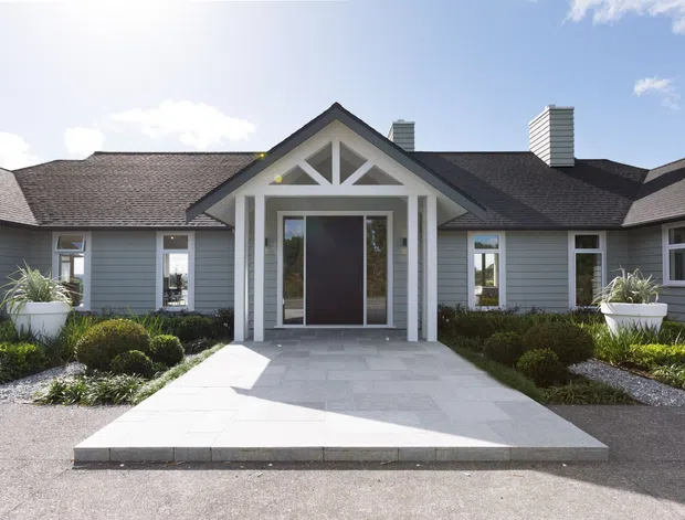 Front exterior view of a broad single-storey home with pale grey horizontal cladding, dark shingle-style pitched roofs, and a prominent gabled entry porch. White timber posts and triangular decorative trusses frame the glazed front door. A paved forecourt and symmetrical low-maintenance landscaping lead to the entrance under a bright blue sky.