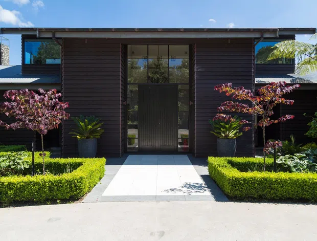 Front exterior view of a contemporary low-rise residence with a dark timber-clad central entry pavilion, tall glazed doorway, broad shallow roof, and symmetrical landscaped garden beds. Pale paving leads to the entrance, with clipped hedges, ornamental trees, and large planters framing the facade.