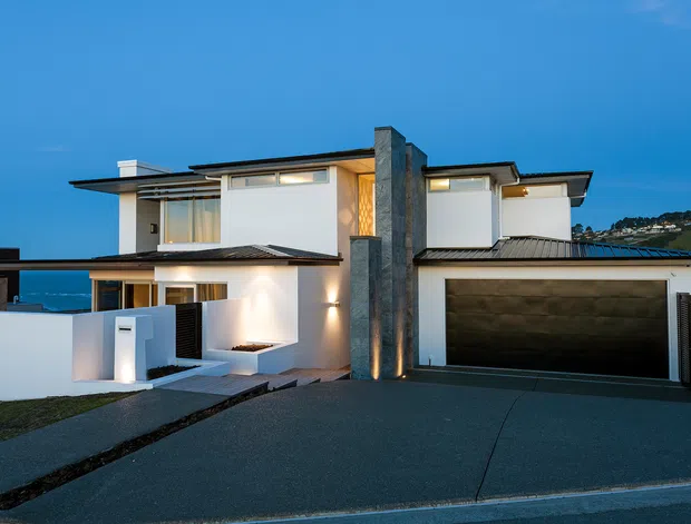 Contemporary multi-level residence photographed at dusk, featuring crisp white rendered walls, dark low-pitched roof planes, extensive glazing, a prominent central stone-clad vertical chimney or feature wall, and an integrated double garage. The home sits on a sloping urban or coastal site with a paved driveway and views toward water and hillside housing in the background.