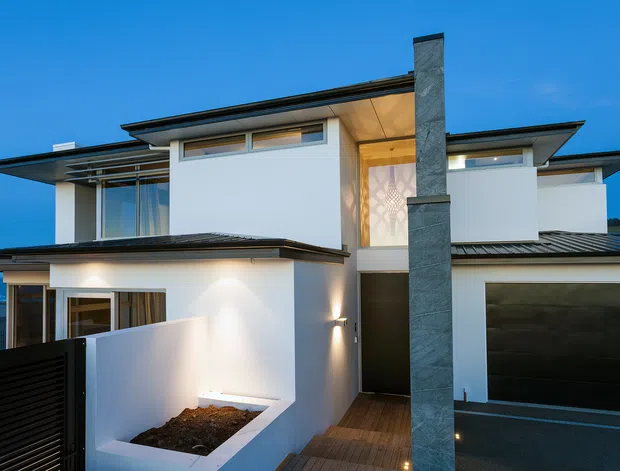 Contemporary multi-level residential home exterior photographed at dusk, featuring crisp white rendered walls, dark metal low-pitched roofs, extensive glazing, a tall stone-clad chimney or vertical feature, timber entry decking, and an integrated dark garage door. The facade has strong horizontal rooflines and clean geometric forms.