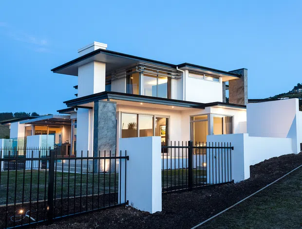 Exterior view of a contemporary two-storey residence at dusk, featuring crisp white rendered walls, dark flat roof planes, extensive glazing, stone-clad vertical elements, and a black metal fence. The home sits on a sloping landscaped site with illuminated interiors visible through the windows.