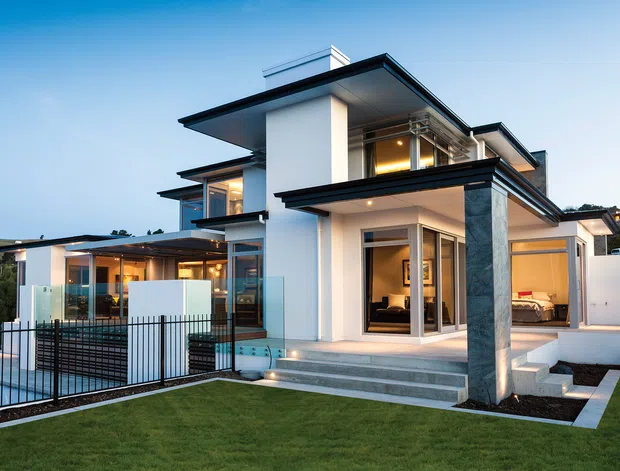 Exterior view of a large contemporary multi-level residence at dusk, featuring crisp white rendered walls, strong horizontal flat roof planes, extensive glazing, dark metal trim, a stone-clad structural column, glass pool fencing, terraces, and landscaped lawn. Warm interior lighting is visible through the windows, with a swimming pool and outdoor living areas in the foreground.