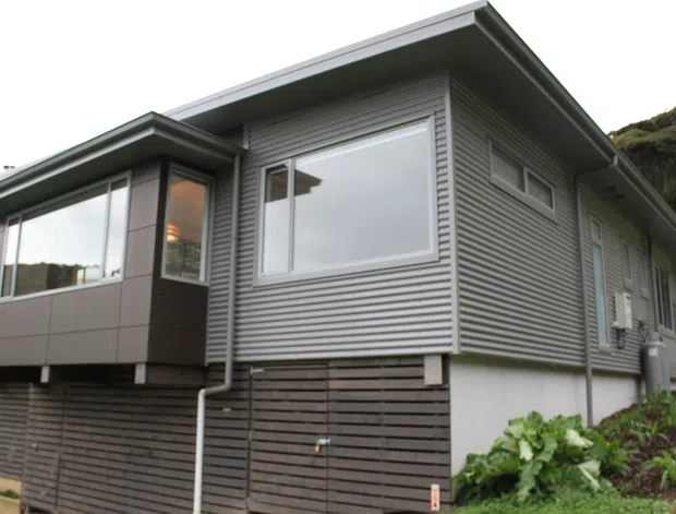 Exterior view of a compact contemporary home on a sloping, leafy site. The building features grey horizontal cladding, dark horizontal timber screening at the lower level, broad overhanging eaves, aluminium-framed windows, and a projecting glazed room or extension.