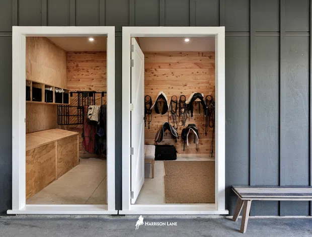 Exterior view of a dark vertical-panelled building facade with two open doorways revealing compact timber-lined storage and changing areas. The left room contains built-in plywood cabinetry and hanging space, while the right room displays multiple hanging items and a small bench. A freestanding bench sits outside along the facade.
