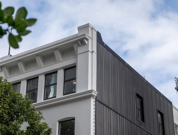 Exterior architectural view of a multi-storey building combining a retained or stylised white classical facade with a contemporary dark vertically ribbed side extension. The facade features projecting cornices, pilasters, decorative brackets and dark-framed windows, while construction activity is visible at the right edge.