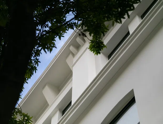 Upward-looking exterior view of a white multi-storey building, framed by leafy tree branches. The facade features smooth rendered walls, strong horizontal moulded bands, recessed dark-framed windows, and projecting geometric architectural elements beneath a pitched or sloping roofline.