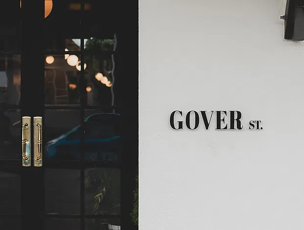 Close-up exterior detail of a commercial or residential entrance, showing black glazed double doors with brass pull handles beside a white rendered wall marked with raised “GOVER ST.” lettering. A wall-mounted exterior light is visible above.