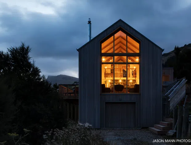 Dusk exterior photograph of a compact, contemporary barn-like dwelling with a steep gabled roof and dark vertical cladding. A large illuminated floor-to-ceiling gable window reveals a warm timber-lined interior, while a garage or storage door occupies the lower facade. The building sits on a sloping, wooded site with exterior stairs and a mountain-like backdrop.