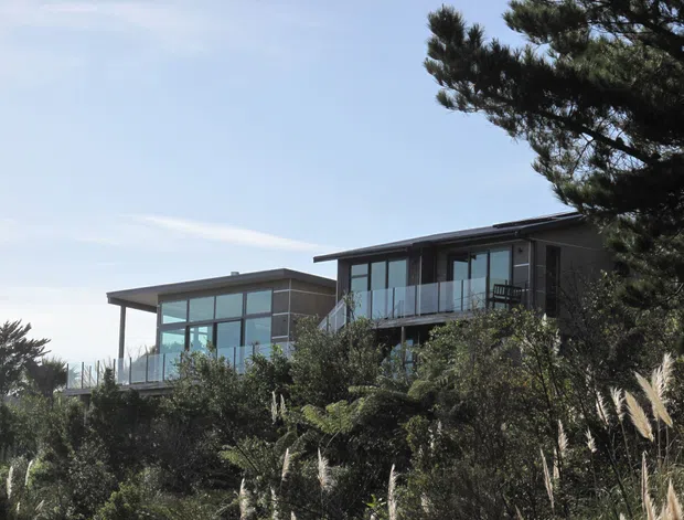 Exterior view of a contemporary multi-level residence set among dense coastal-style vegetation. The house features flat, low-pitched roof planes, extensive floor-to-ceiling glazing, dark exterior cladding, and glass balcony balustrades.