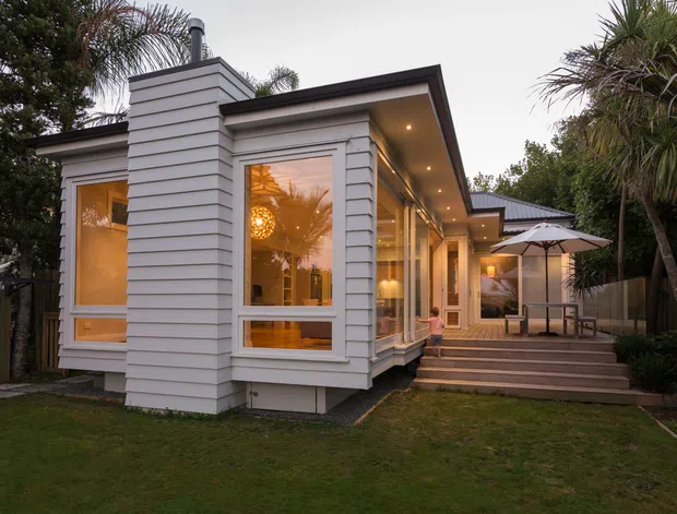 Exterior view of a single-storey residential extension or bungalow-style home, with white horizontal weatherboard cladding, large timber-framed windows, a projecting chimney volume, and a covered outdoor deck accessed by broad steps. Warm interior lighting glows through the glazing, with outdoor dining furniture and an umbrella on the deck. The property is surrounded by established tropical and subtropical planting and lawn.