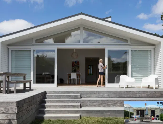 Front exterior view of a bright, single-storey contemporary gable-roof house with a broad timber deck and stepped entry. The facade features extensive floor-to-ceiling glazing, white horizontal cladding, a triangular clerestory window, and deep roof eaves. Outdoor seating and a picnic-style table occupy the deck, while a small inset image at lower right shows the previous condition of the facade.