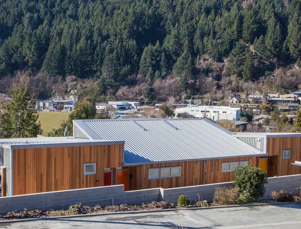 Exterior view of a contemporary low-rise building with vertical timber-clad wall sections, light corrugated metal pitched roofing, dark masonry boundary walls, and modern windows. The building sits in a suburban setting against a densely forested hillside.