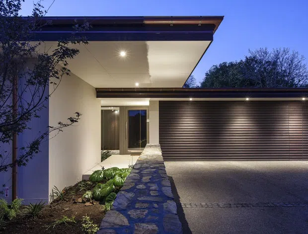 Dusk exterior view of a modern single-storey residence, featuring a broad cantilevered flat roof, recessed entry, dark horizontal garage door, smooth light walls, and a stone-paved path and retaining wall. Exterior lighting highlights the entry and driveway, with landscaped planting along the approach.