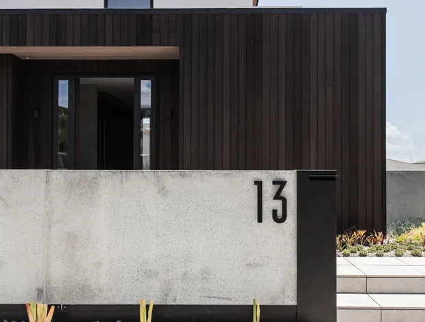 Front exterior view of a contemporary two-storey residence with a minimalist rectilinear form. The upper level has smooth light plaster walls and a narrow dark-framed window, while the lower facade is clad in dark vertical timber boards around a recessed entry. A pale concrete boundary wall displays the number 13 beside a dark mailbox structure, with broad concrete steps, gravel landscaping, and sparse architectural planting leading to the home.