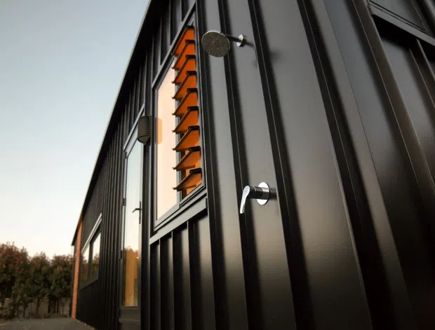 Close-up exterior view of a contemporary building clad in dark vertical metal panels, featuring tall windows with prominent orange louvres and modern exterior wall-mounted lighting. The image emphasizes the facade detailing and strong vertical composition.