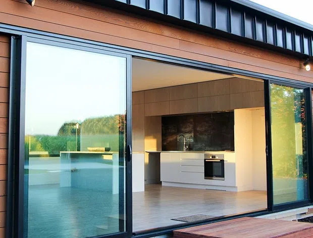 Exterior view of a contemporary single-storey dwelling with warm horizontal timber cladding and dark metal trim. Large sliding glass doors open onto a timber deck, revealing a minimalist kitchen with white cabinetry, integrated oven, pale flooring, and a dark stone or tiled splashback. A pool or reflective water feature is visible beyond the glazing.