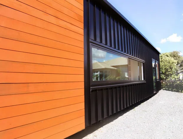 Exterior view of a contemporary building with contrasting orange timber horizontal cladding and dark vertically ribbed wall cladding. A large recessed window reflects a light-colored structure, while a gravel path runs alongside the facade. Bright sunny conditions and neighboring residential elements are visible in the background.