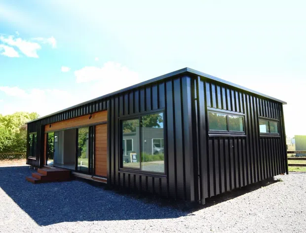 Exterior view of a compact single-storey modern building with black vertically ribbed metal cladding, large windows, and a warm timber-clad entrance section. The structure has a simple low-pitched roof and sits on a gravel-covered site.