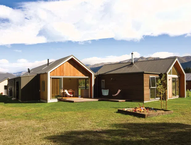 A single-storey modern cabin-style residential building composed of connected dark-clad volumes with prominent timber-lined gable ends, large glazed doors, and a broad lawn. Mountain ranges are visible in the background under a bright sky, and a small raised garden bed sits in the foreground.