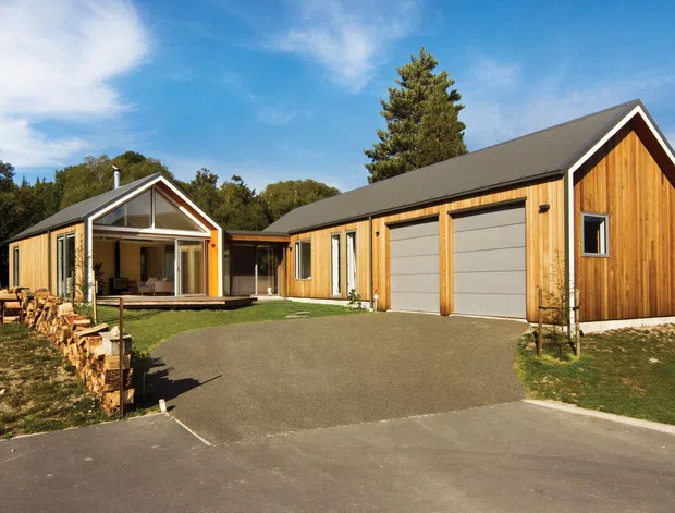 Exterior view of a contemporary timber-clad, barn-inspired residential building composed of a main garage/workshop wing and a smaller gabled living pavilion. The structures feature dark pitched roofs, natural vertical timber weatherboarding, white-trimmed openings, large glazed doors, and a broad paved driveway in a wooded setting.