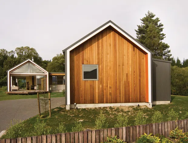 Exterior view of a compact modern gable-roof building clad in warm vertical timber boards, with contrasting dark metal side cladding and white trim. A glazed pavilion-like structure is visible behind it, set within a landscaped suburban or rural garden.