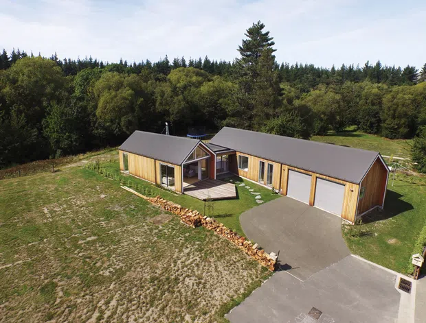 Aerial view of a modern single-storey timber-clad residence arranged around a small central courtyard or covered entry deck. The home has multiple pitched gable roof volumes, a large attached double garage, broad glazing, and a concrete driveway, set within a spacious grassy and wooded rural landscape.