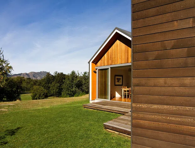 Exterior view of a small timber-clad gabled building with a glazed sliding door opening onto a timber deck. A larger dark timber-clad wall occupies the foreground, with lawn, trees and mountain terrain in the background.