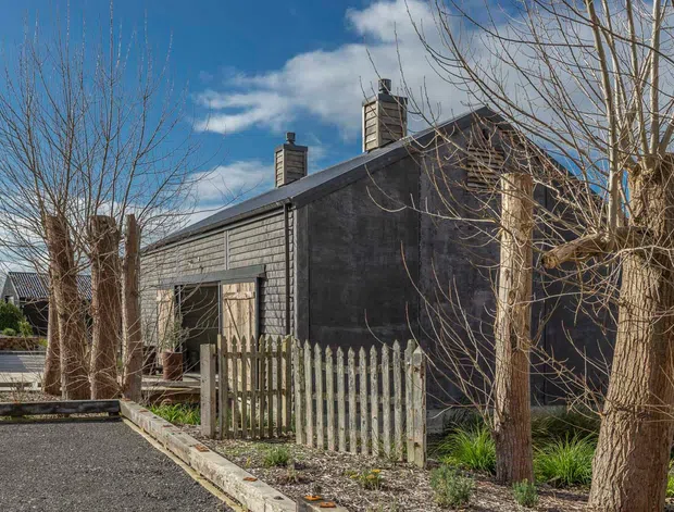 Exterior view of a dark, weathered-clad single-storey building with a pitched roof, multiple chimneys, timber fencing, and a paved driveway or walkway. Leafless trees frame the property, with neighbouring structures partially visible in the background.