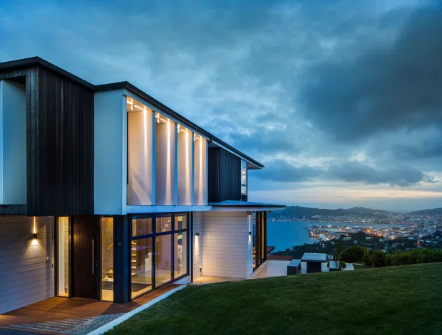 Twilight exterior view of a contemporary multi-level house on a sloping site, with dramatic angular roof forms, dark vertical cladding, light horizontal wall cladding, extensive glazed openings, and warm interior lighting. A landscaped lawn and expansive coastal urban bay panorama are visible beyond the building.