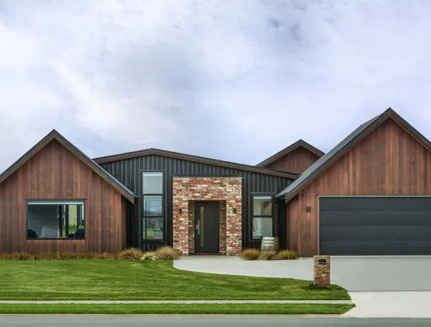 Front exterior view of a contemporary single-storey home with multiple steep gabled roof forms, dark vertical metal cladding, warm timber gable accents, a central recycled-brick entry surround, and an integrated double garage. The property has a manicured lawn, concrete path and driveway, with subdued mountain or rural landscape visible in the background.