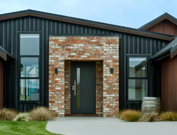 Front exterior view of a contemporary single-storey home with dark vertical metal cladding, a prominent weathered red-and-white brick entry surround, dark-framed glazing, and intersecting pitched roof forms. Ornamental grasses and a curved concrete path frame the entrance.
