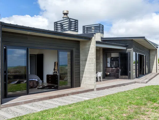 Exterior view of a low, contemporary single-storey home with dark horizontal cladding, pale masonry chimney-like walls, broad glazed sliding doors, covered outdoor living areas, and flat-pitched roof sections. A paved terrace and lawn extend along the facade, with rooftop screened structures and a visible flue.