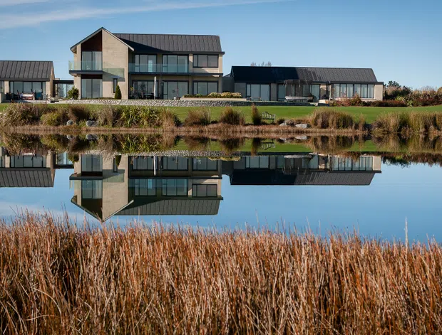 Wide exterior view of a contemporary multi-building residence beside a calm pond, with strong reflections in the water. The main house has a steep gabled roof, dark metal roofing, extensive glazing, and a mix of dark cladding and pale masonry or rendered walls. Low wing buildings extend on both sides, with landscaped grasses and reeds in the foreground and a clear blue sky.