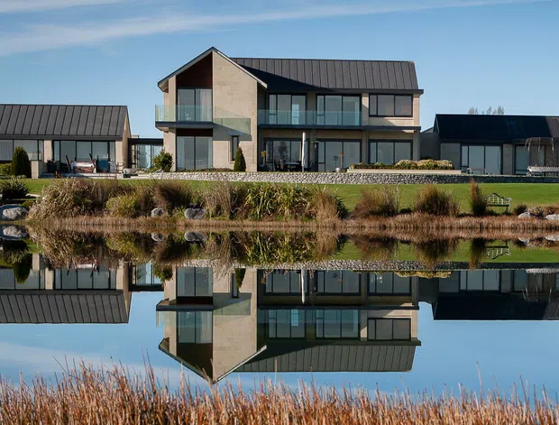 A wide exterior view of a contemporary multi-building residence beside a landscaped pond. The central two-storey house has a steep gabled roof, extensive dark-framed glazing and a pale rendered facade, flanked by two long, low outbuildings with dark metal pitched roofs. The buildings and surrounding reeds are reflected clearly in the still water.