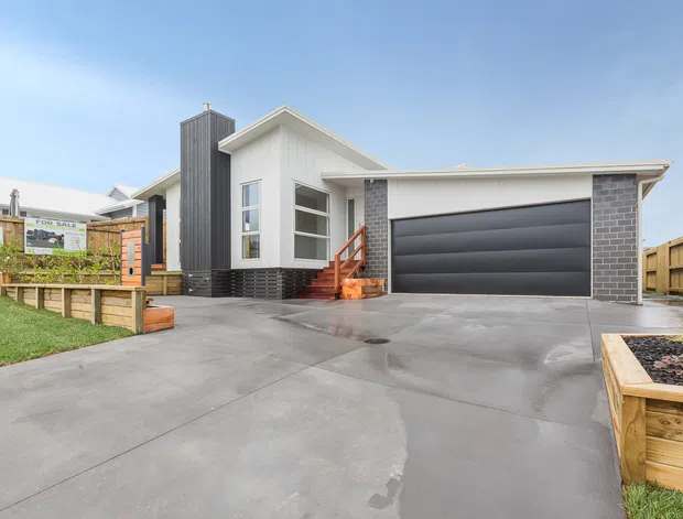 Street-facing exterior view of a contemporary single-storey home with a gabled roof, white vertical cladding, dark brick feature walls, a tall charcoal-clad entry volume, and an attached double garage with a dark sectional door. A broad concrete driveway, timber fencing, raised garden beds, and front steps are visible.