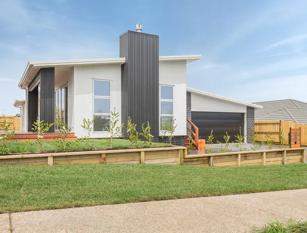 A contemporary single-storey suburban house viewed from the street, featuring white rendered walls, a prominent dark vertical-clad chimney or entry volume, grey metal roofing, large windows, and an attached covered carport. The property has timber fencing, raised timber garden edging, and a landscaped front garden.
