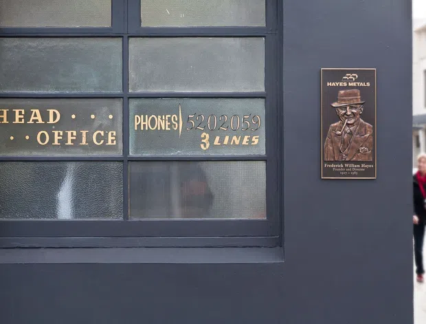 Close-up of a dark painted commercial or industrial-style facade featuring a large multi-pane textured-glass window. Gold lettering on the glass reads “HEAD OFFICE” and “PHONES 5202059 3 LINES,” while a vintage-style Hayes Metals plaque is mounted on the wall beside it. A blurred pedestrian and neighboring building are visible in the background.