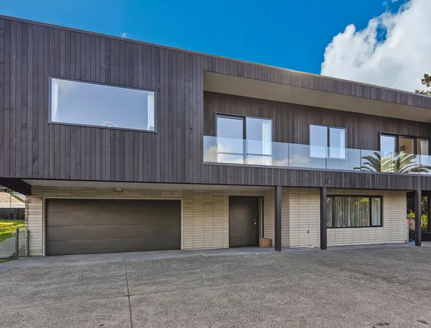Exterior view of a contemporary two-storey residence with a dark vertical timber-clad upper level, pale masonry lower level, integrated garage, recessed entry, and a long recessed balcony with glass balustrade. The home has a clean, rectilinear form and a low-pitched roofline, set against a bright blue sky.