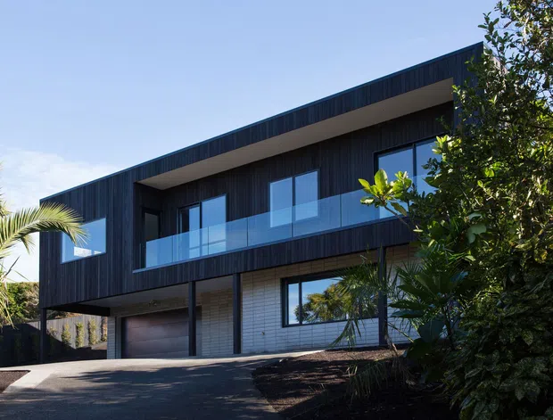Contemporary two-storey residence with a bold dark-clad upper level, recessed balcony with clear glass balustrade, large rectangular windows, and a pale brick or masonry ground floor. A wide garage door and paved driveway occupy the lower level, while established tropical and subtropical landscaping frames the facade.