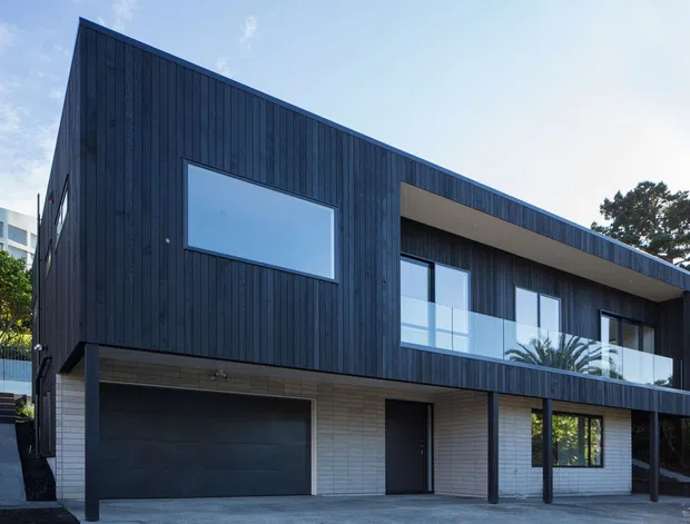Exterior view of a contemporary two-storey residence with a steep asymmetric roofline, dark vertical timber cladding, pale brickwork at ground level, broad glazing, a recessed upper balcony, and an integrated dark garage door. The home sits in a leafy suburban setting.