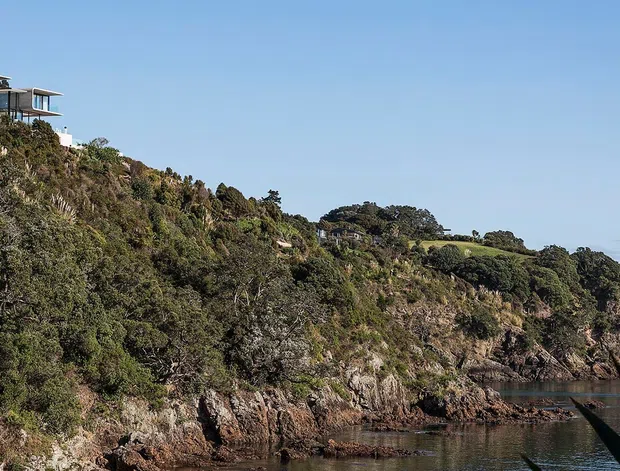 A wide exterior landscape view of a modern home perched on a steep rocky, vegetated coastal or waterfront cliff. The building has expansive glazing, slender structural elements, and a low-pitched flat roof form, with calm water and rugged shoreline below.