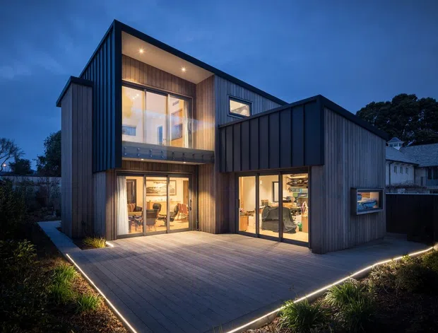 Dusk exterior view of a contemporary multi-level home with dark vertical cladding, angular projecting volumes, extensive glazed doors and windows, and a large timber-toned deck edged with integrated linear lighting. Warm interior lighting reveals living spaces inside, while landscaped garden beds frame the foreground.