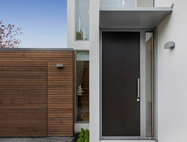 Modern residential entry facade featuring a tall dark front door recessed beneath a projecting upper-level structure, set within smooth white walls. A horizontal timber-clad garage or privacy door occupies the left side, with narrow glazing and minimalist exterior lighting adding to the clean contemporary composition.